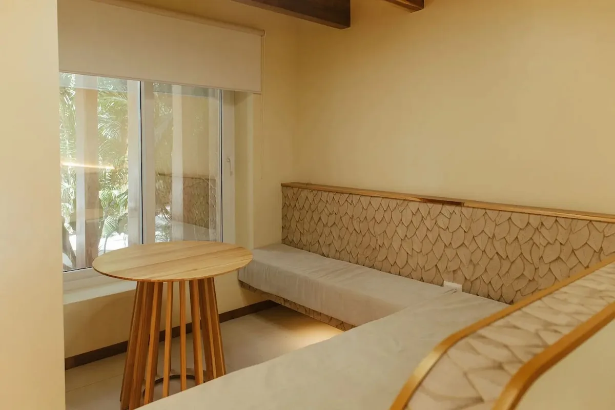 Dormitorio en tonos claros con una cama parcialmente visible, banco tapizado en forma de L, mesa redonda de madera, ventana y vigas de madera en el techo.