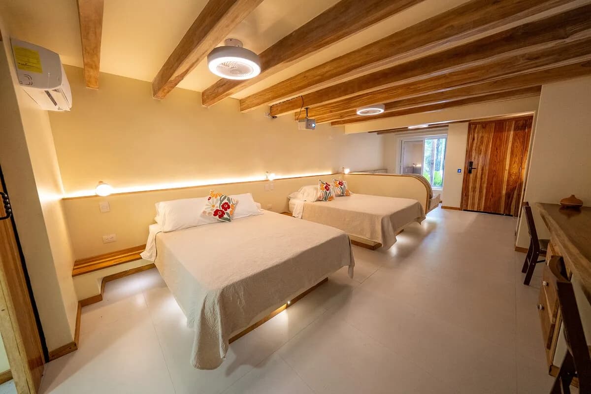 Dos camas vestidas de blanco bajo vigas de madera, con mesas pequeñas, escritorio lateral y puerta de madera al fondo.