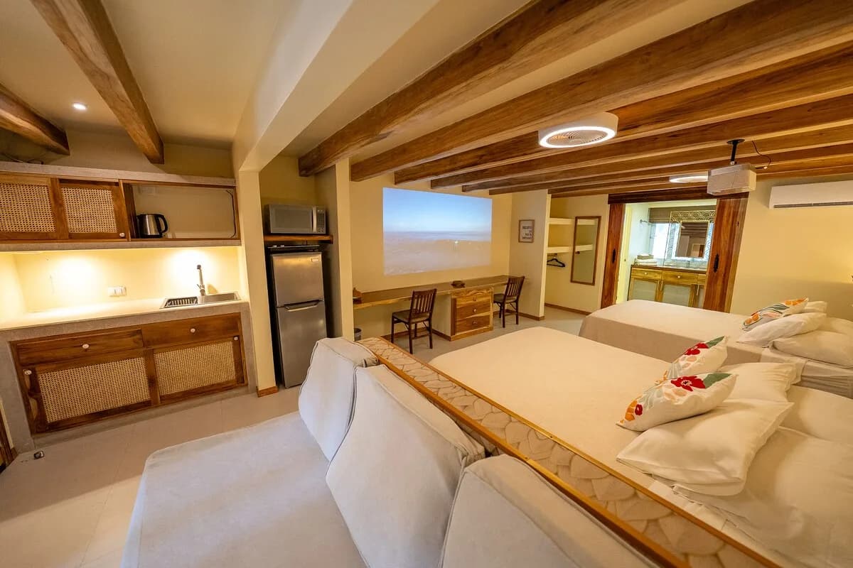 Dormitorio con dos camas, mueble con fregadero y refrigerador a la izquierda, escritorios con sillas y una imagen rectangular proyectada en la pared.