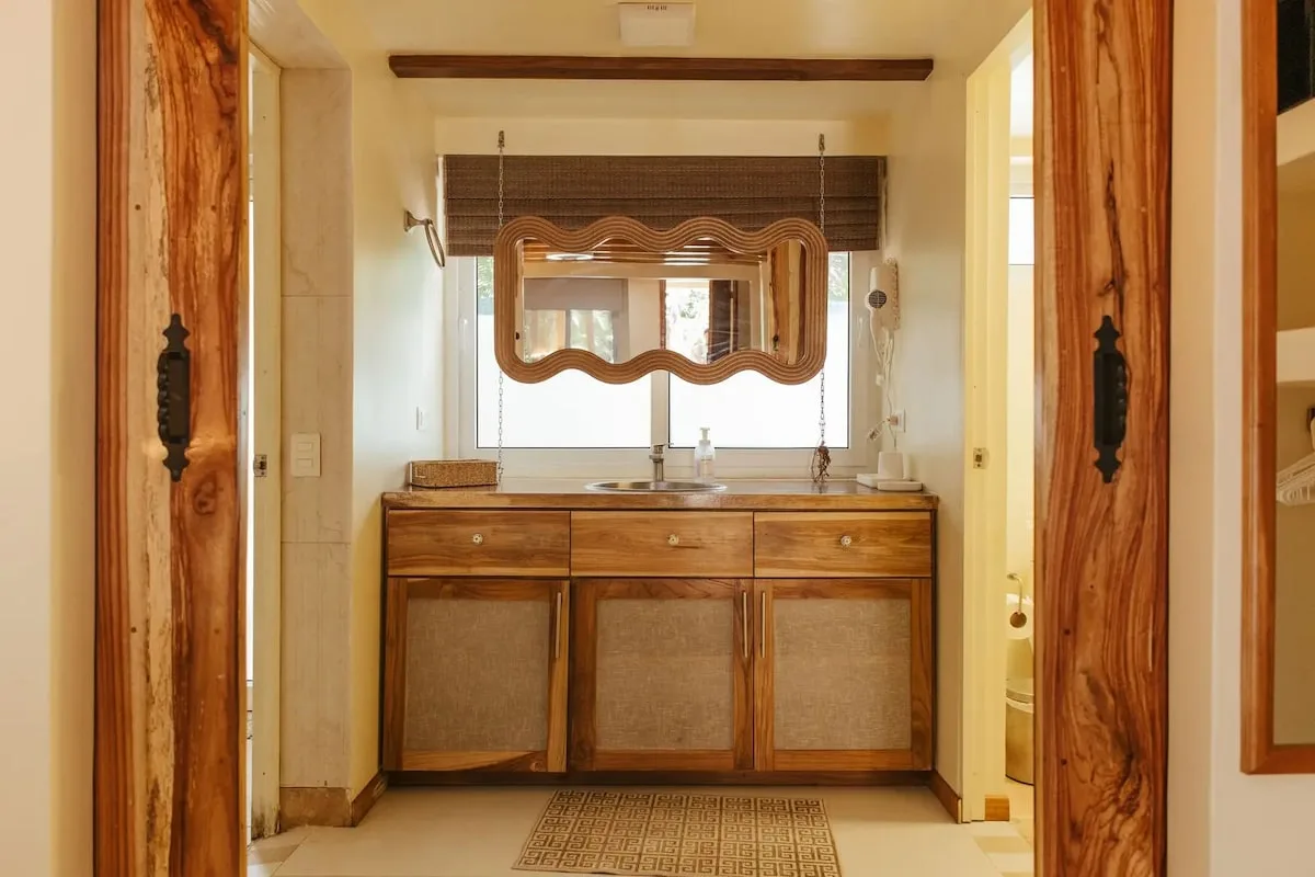 Mueble de lavabo de madera visto entre dos puertas, con pila redonda, grifo, espejo de marco ondulado y persiana oscura.