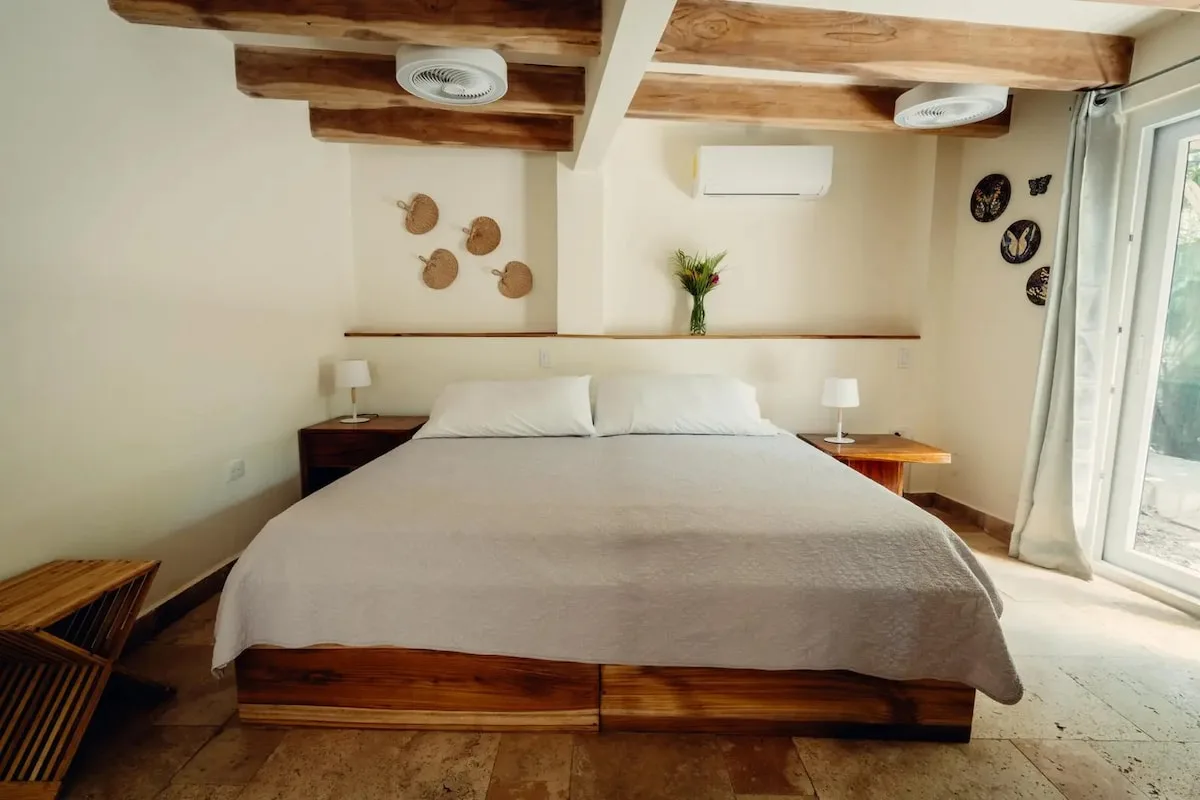 Cama grande centrada entre dos mesas con lámparas, bajo vigas de madera; una puerta acristalada ocupa el lado derecho.