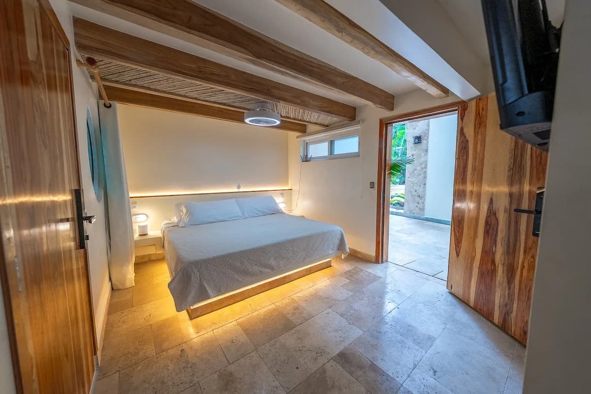 Dormitorio visto desde la entrada, con una cama centrada, vigas y entramado de madera en el techo y puertas abiertas hacia un paso exterior.