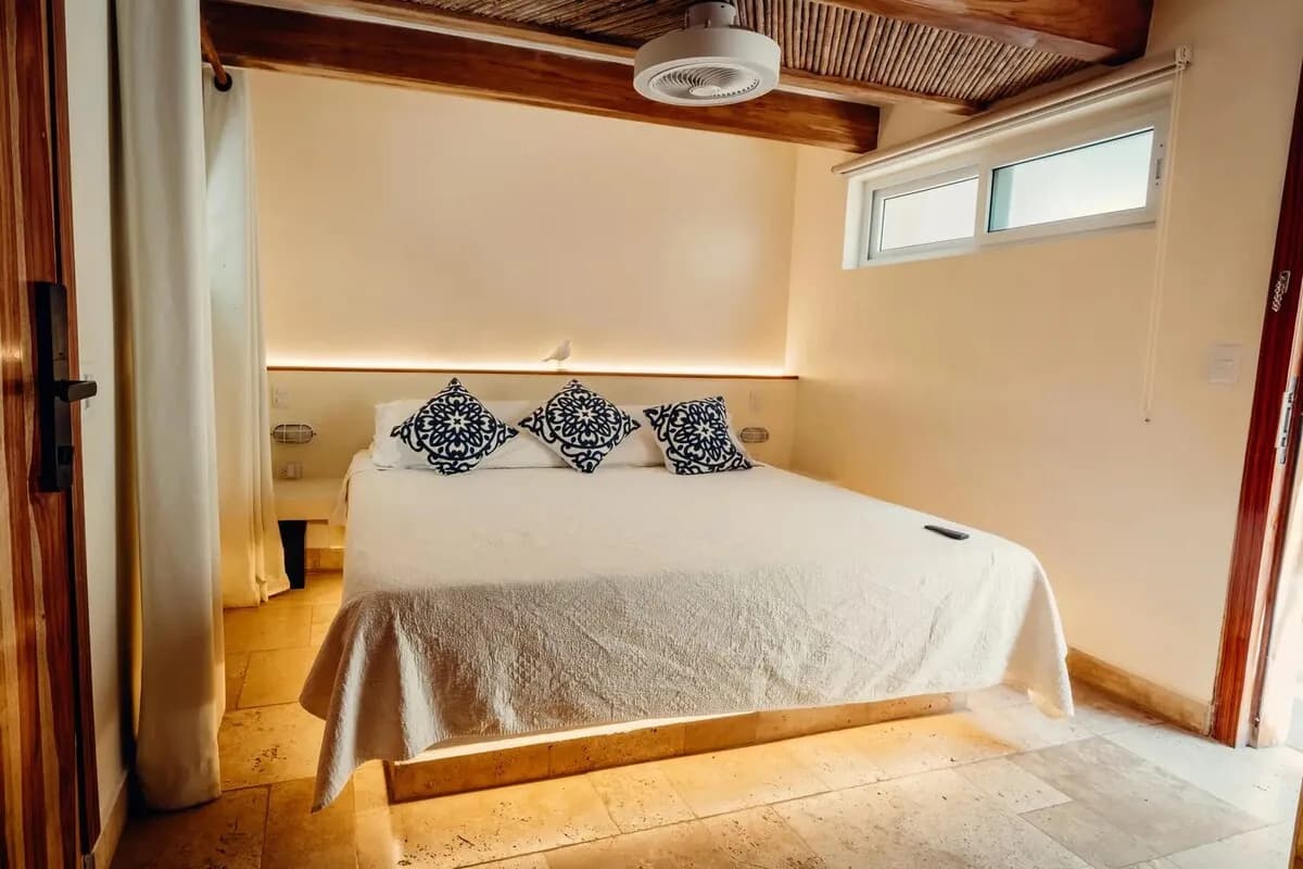 Cama con tres cojines estampados bajo vigas de madera y techo entramado, con ventana horizontal alta en la pared.