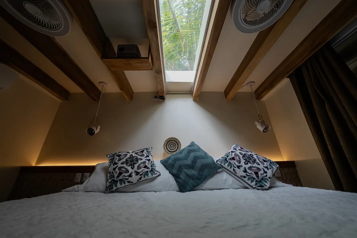 Vista baja y cercana de una cama con cojines estampados, bajo vigas de madera y una abertura acristalada larga en el techo.