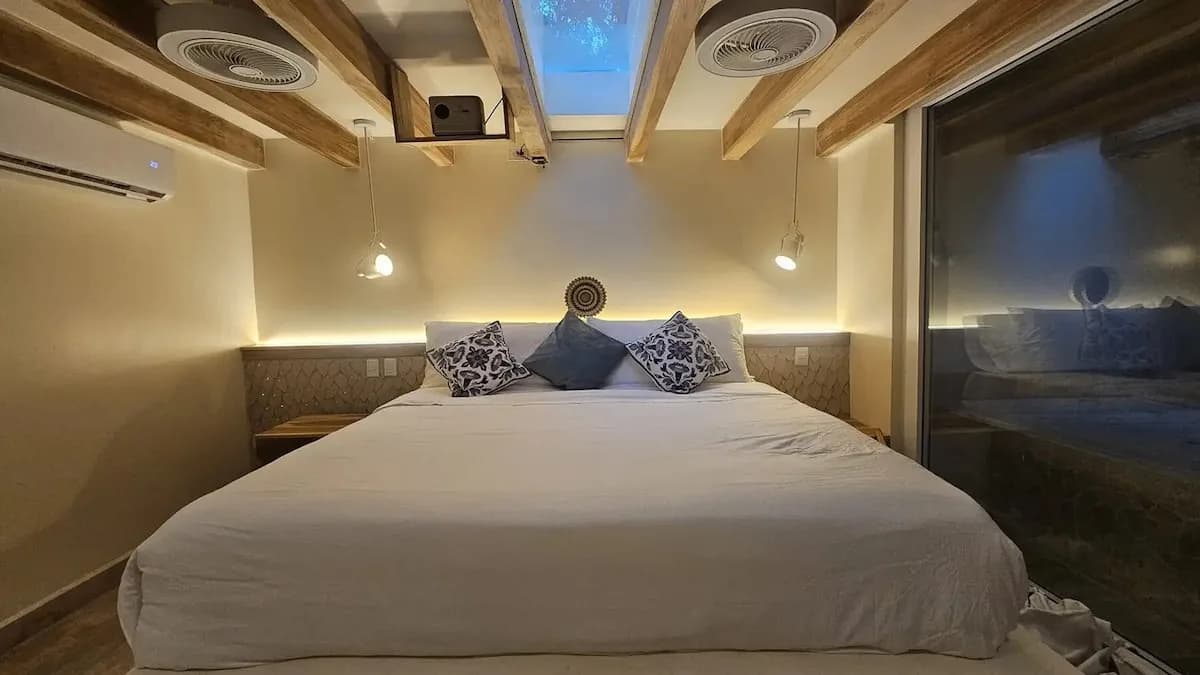 White bed centered beneath wooden beams, a skylight, and pendant lights, with a large dark glass panel on the right.
