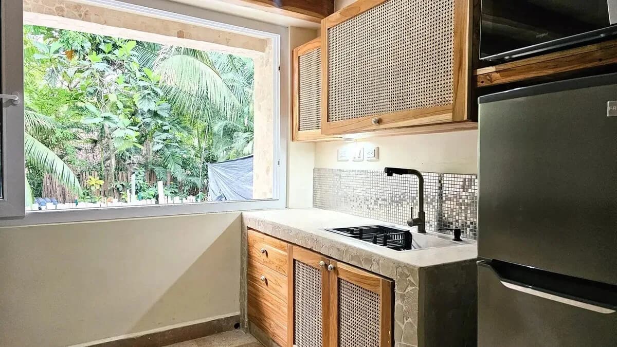 Compact kitchen with wood-and-cane cabinets, a black sink, a silver refrigerator, and an open window facing dense greenery.