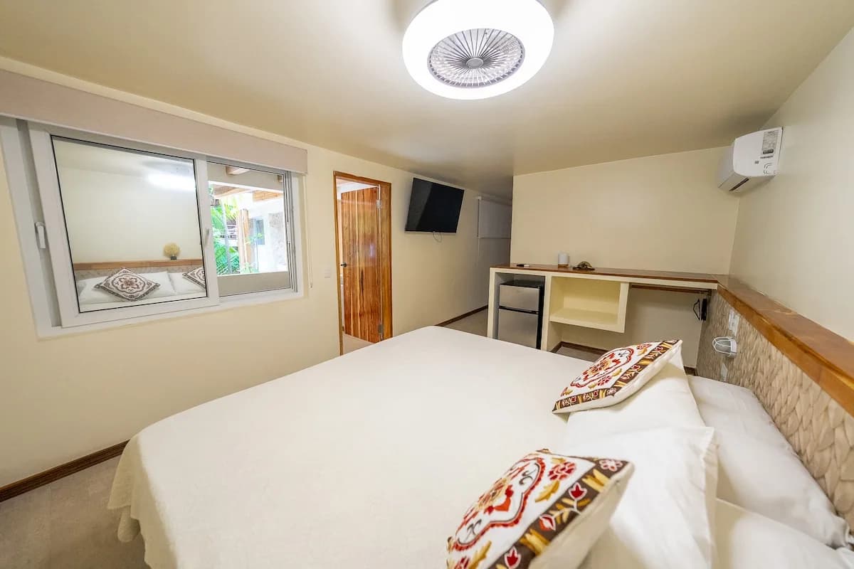 Vista amplia sobre una cama blanca hacia una ventana interior, puerta de madera, pantalla mural y barra con refrigerador y estantes.
