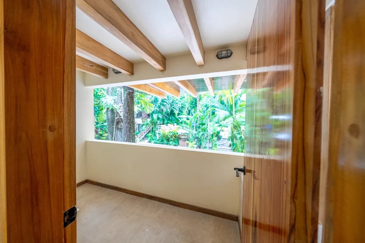 Corredor cubierto vacío con vigas de madera, puertas abiertas a ambos lados y una abertura amplia hacia vegetación tropical.