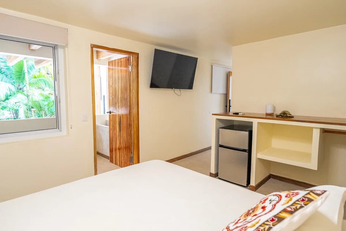 Dormitorio con cama en primer plano, ventana hacia palmeras, puerta abierta, pantalla mural y refrigerador bajo una barra.