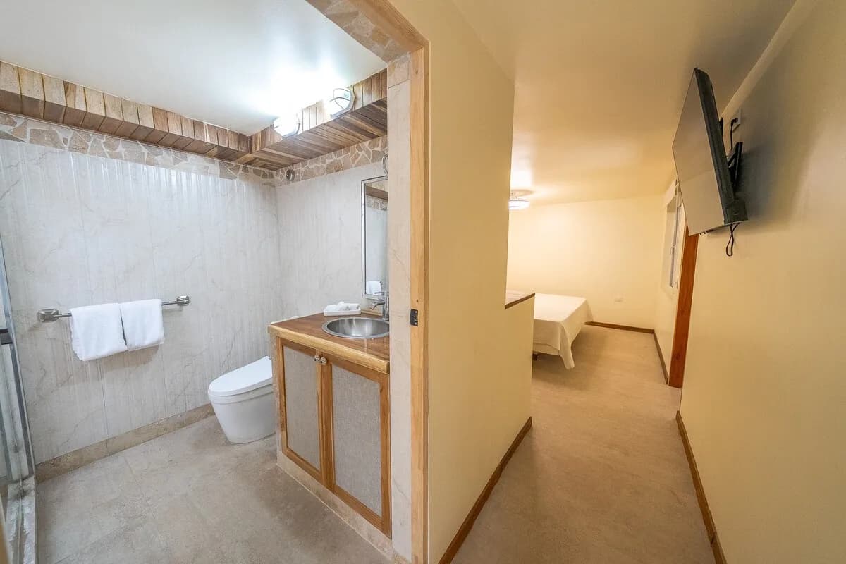 Composición dividida entre un baño con inodoro y lavabo a la izquierda y un pasillo que conduce a una cama al fondo a la derecha.