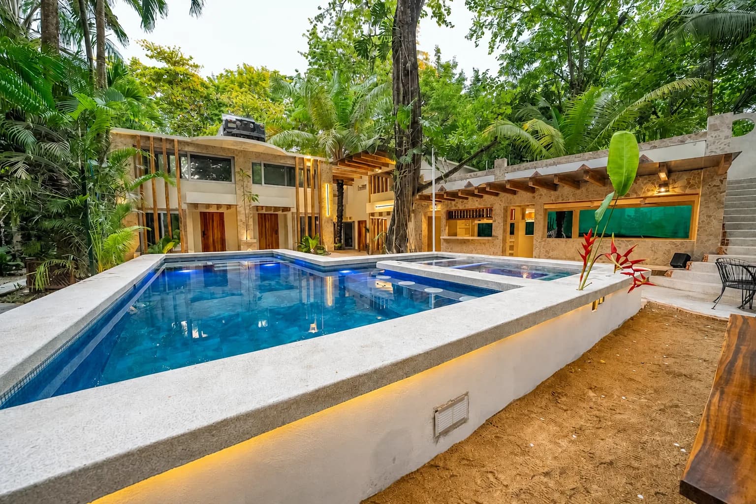 Gran piscina azul en primer plano, rodeada por edificios revestidos de piedra, árboles tropicales y una zona de mesas a la derecha.
