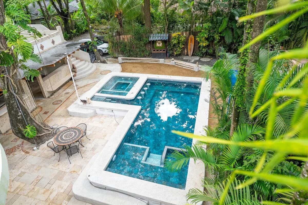 Vista elevada de una piscina azul de forma angular, patio de piedra con mesa redonda, zona de arena y vegetación tropical alrededor.