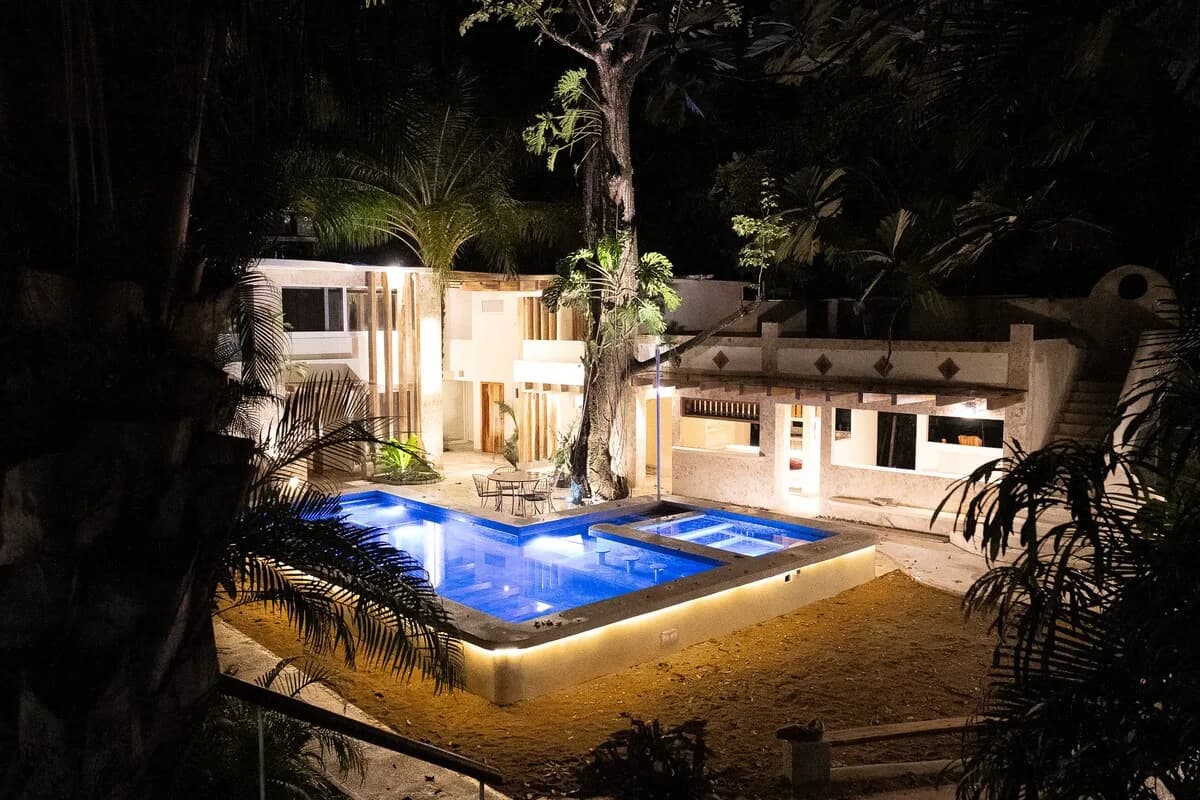 Vista nocturna elevada de piscinas iluminadas en azul y edificios claros, enmarcados por troncos y hojas oscuras.