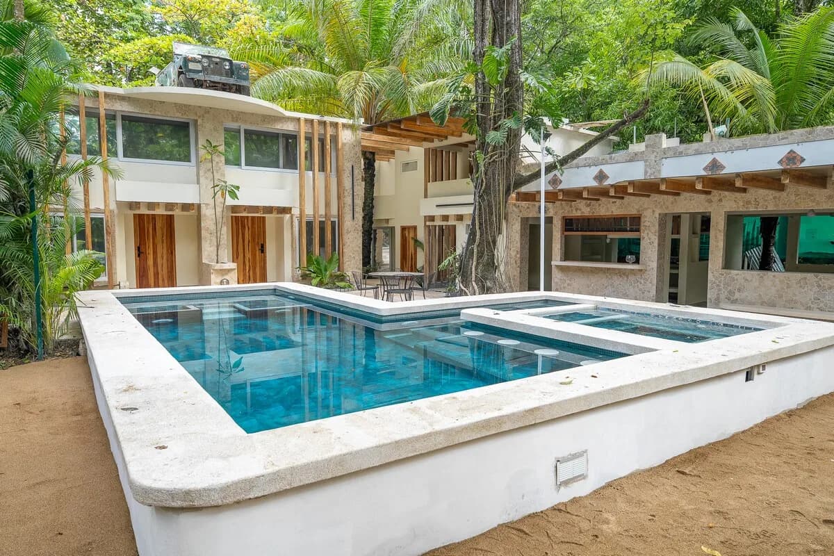 Piscina azul de bordes blancos frente a dos edificios revestidos de piedra, con un gran árbol en el centro y vegetación densa alrededor.
