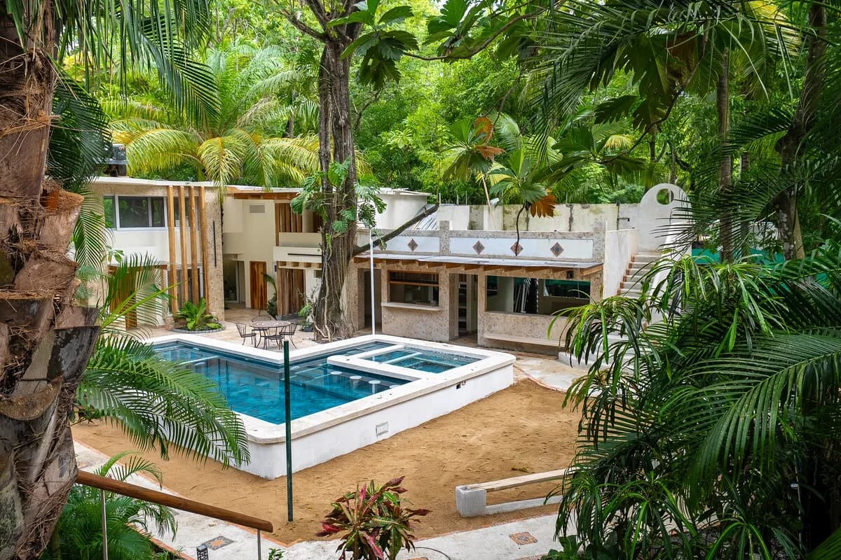 Vista elevada a través de palmeras hacia una piscina, una zona de arena, edificios de piedra y una escalera exterior.